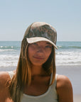 Woman wearing a leopard camo cap with 'ARIKO' branding on a beach
