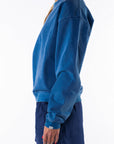 Side view of a woman wearing a blue post surf crew sweatshirt and navy day shorts on a white background