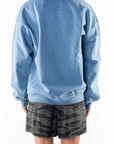 Back view of a woman wearing a teal sweatshirt and ocean tiger camo day shorts on a white background