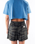 Back view of a woman wearing a teal crop t-shirt and ocean tiger camo day shorts on a white background