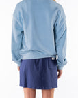 Back view of a woman wearing a light blue ocean sweatshirt and navy day skirt on a white background