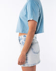 Side view of a woman wearing a turquoise beach crop t-shirt with a denim mini skirt on a white background.