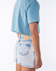 Side back view of a woman wearing a turquoise beach crop t-shirt with a denim mini skirt on a white background.