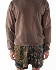 Back view of a person wearing a brown sweatshirt and real tree camouflage day shorts on a white background