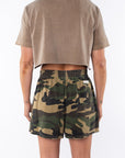 Back view of a woman wearing a brown beach crop t-shirt and camouflage day shorts on a white background