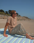 Person sitting on a striped towel at the beach with a clear sky. He is wearing beach t-shirt in brown and leopard camo day pants.