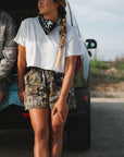 woman leaning against truck wearing real tree camo day shorts, crop tee and beach bandana
