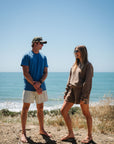 Two people standing on a beach with clear blue sky and ocean. Man is wearing blue beach t-shirt and beige french terry day shorts and ocean tiger surf trucker hat. She is wearing brown french terry post surf crew sweatshirt and day shorts.