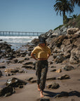 woman walking on the beach. She is wearing beach crop tee in sunrise yellow with real tree camo day pants and 'ARIKO' logo surf trucker hat.