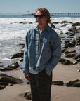 man wearing denim jacket with ocean tiger camo day pants, sunglasses at the beach