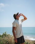 Man drinking a soda by the ocean on a clear day. He is wearing a beige beach t-shirt and french terry day shorts in dark gray.