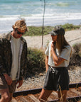 Two people walking along a coastal path with ocean in the background. Man is wearing a real tree camo bucky jacket and brown french terry day shorts. She is wearing a beach crop t-shirt in beige with dark gray french terry day shorts and an ocean tiger camo surf trucker.
