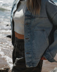 close up of woman wearing denim jacket, beach crop tee in sandstone and ocean tiger day shorts at the beach