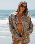 Woman on a beach wearing sunglasses and a bucky leopard camo patterned jacket, holding flowers.
