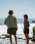 Two people standing on a rocky beach wearing ARIKO clothing with ocean and sky in the background