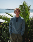 man against palm leaves wearing teal post surf crew sweatshirt and army green day shorts.