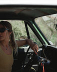 woman in a truck looking back. she is wearing the surf tank in sunrise yellow. There is a 'ARIKO' logo surf trucker hat on the dashboard.