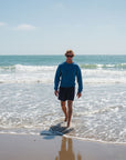 Man walking on a beach with ocean waves in the background. Blue post surf crew sweatshirt and navy day shorts.