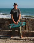 Man holding a teal skateboard on a beach with ocean waves in the background. He is wearing a 'KOOK' graphic t-shirt with army green day pants and an ocean tiger surf trucker hat.