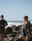 two models standing on the beach. male is wearing post surf crew sweatshirt in sea grey with army green day shorts. female is wearing summer softy plaid shirt in black/red with beach crop tee in sea grey and navy day shorts.