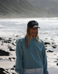 woman wearing post surf crew sweatshirt in ocean with 'ARIKO' OG logo surf trucker hat