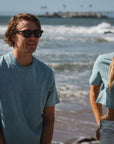 man and woman wearing beach t-shirt and beach crop tee in ocean. woman has OG ARIKO Logo surf trucker hat on.