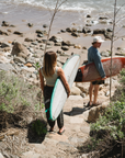 Two people carrying surfboards walking up a rocky path towards the ocean. Wearing ARIKO clothing.