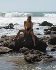 woman sitting on a rock in the ocean. she is wearing the scallop tank in sunrise yellow and camo day shorts