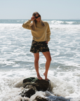 woman on a rock in shallow ocean wearing post surf crew sweatshirt in sunrise yellow with camo day shorts