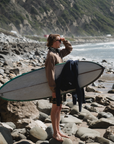 man wearing post surf crew sweatshirt in brownstone while holding a surfboard looking at the waves.