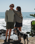 woman facing away towards beach wearing summer softy plaid shirt in green/orange with navy day shorts. man next to her is wearing post surf crew sweatshirt in sandstone and army green day shorts.