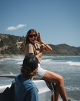woman sitting on a truck wearing beach crop tee in brownstone with denim day skirt laughing with a man wearing beach t-shirt in teal. Both at the beach.