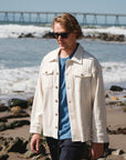 Man wearing a Beige Bucky Shirt Jacket and sunglasses on a beach with waves and a bridge in the background