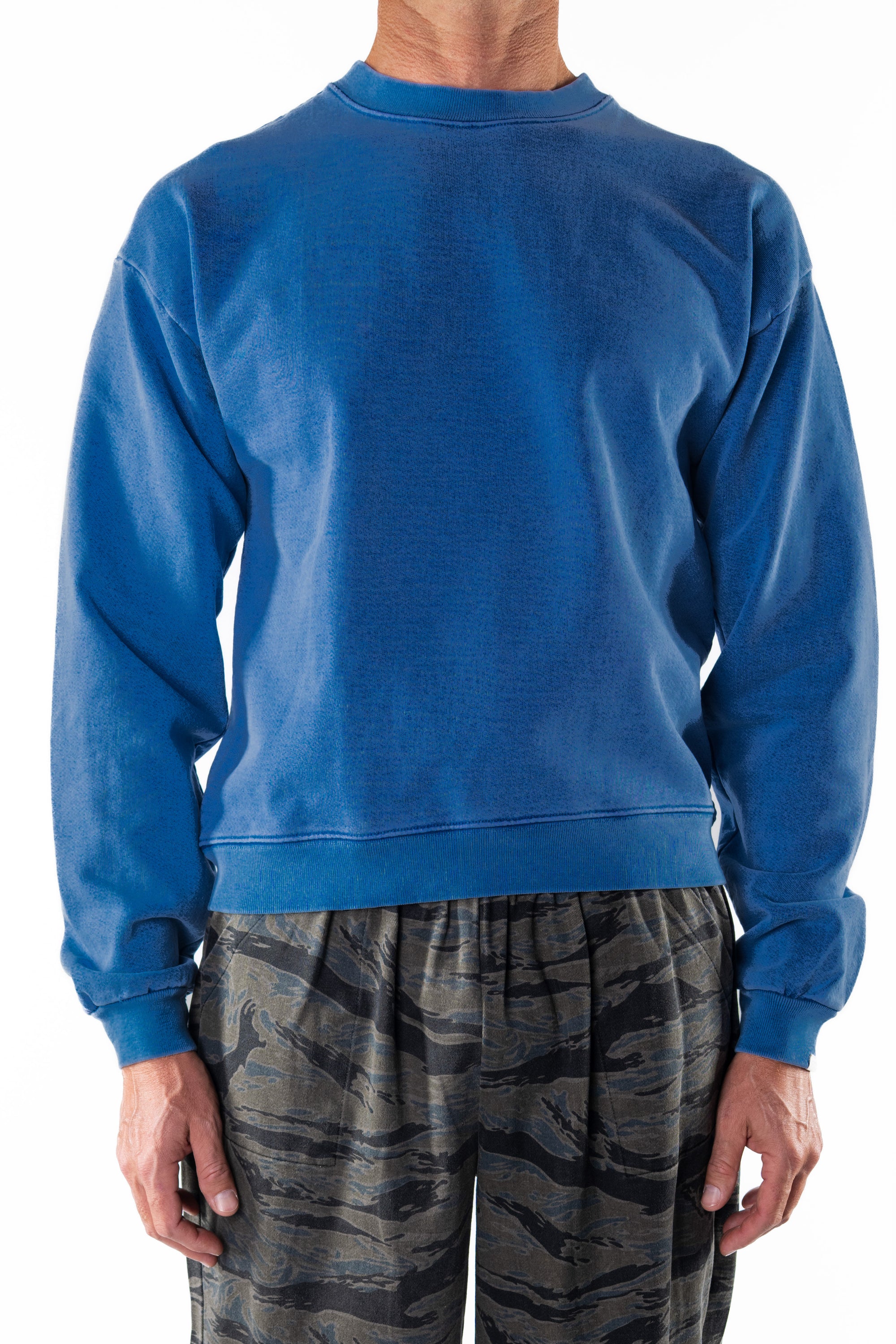 Person wearing a blue post surf crew sweatshirt and ocean tiger camouflage day pants on a white background