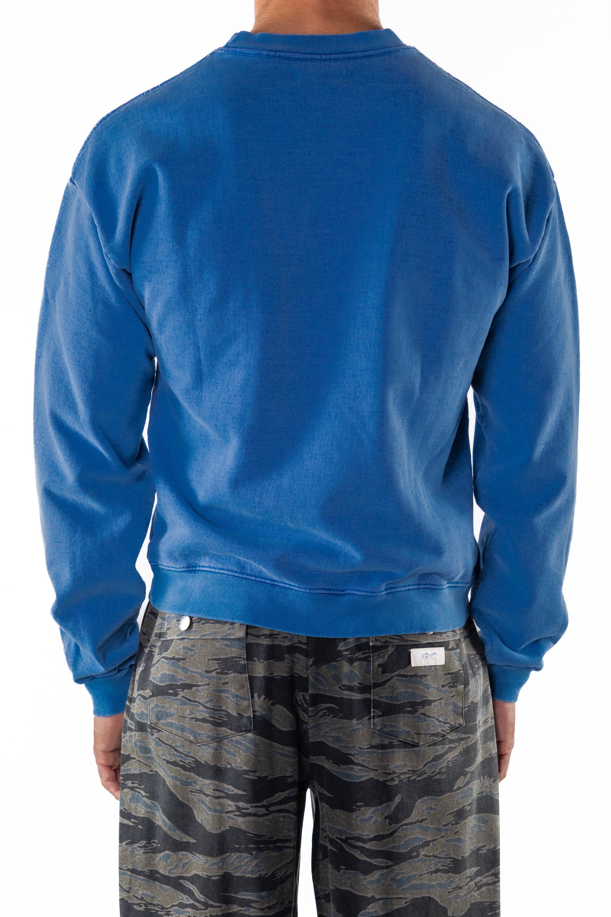 Back view of a person wearing a blue post surf crew sweatshirt and ocean tiger camouflage day pants on a white background