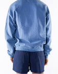 Back view of a woman wearing a blue sweatshirt and navy shorts on a white background