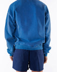 Back view of a woman wearing a blue post surf crew sweatshirt and navy day shorts on a white background