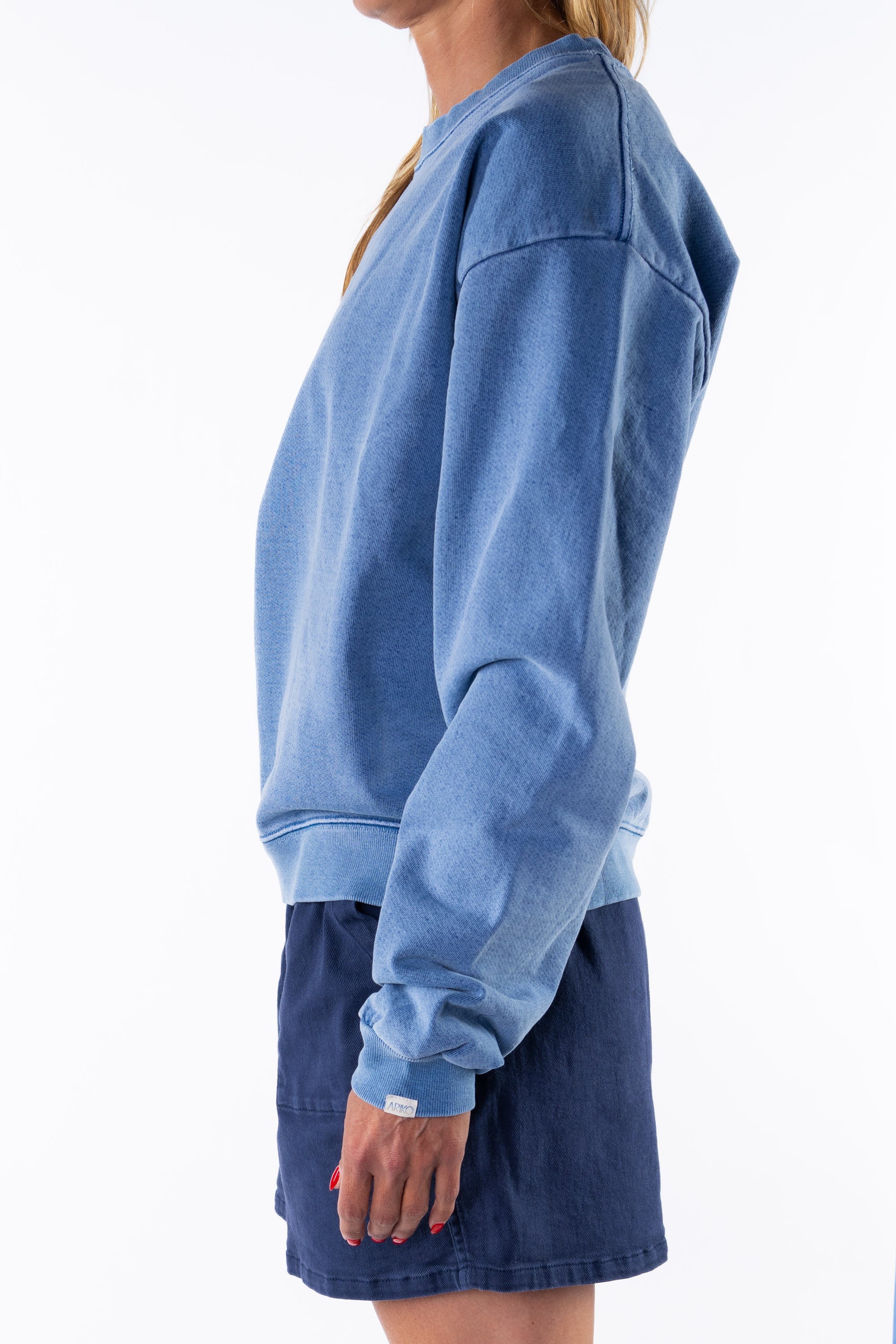 Side view of a woman wearing a blue sweatshirt and navy day shorts on a white background