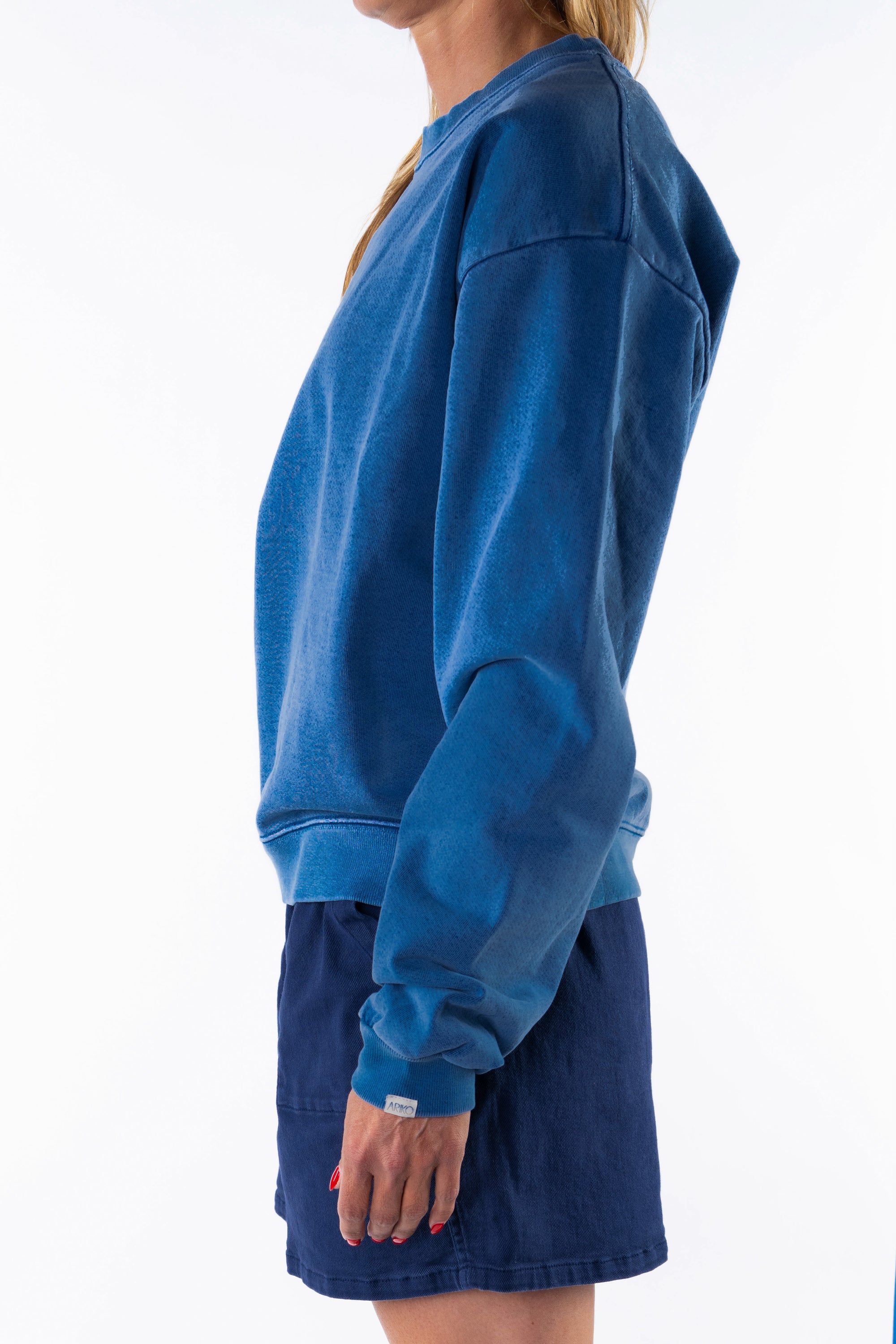 Side view of a woman wearing a blue post surf crew sweatshirt and navy day shorts on a white background