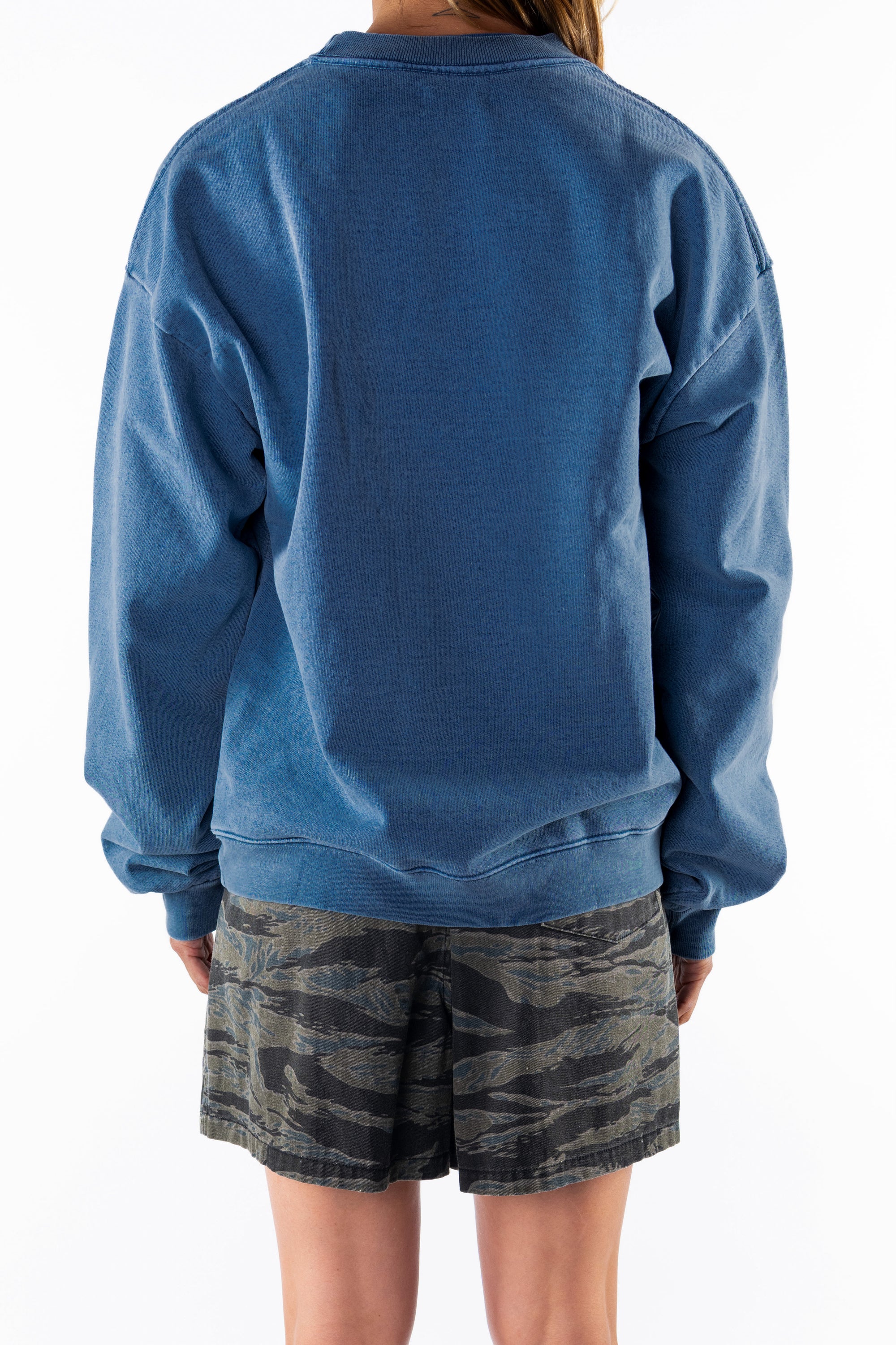 Back view of a woman wearing a teal post surf crew sweatshirt and ocean tiger camouflage day shorts on a white background