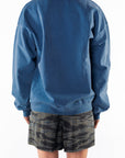 Back view of a woman wearing a teal post surf crew sweatshirt and ocean tiger camouflage day shorts on a white background