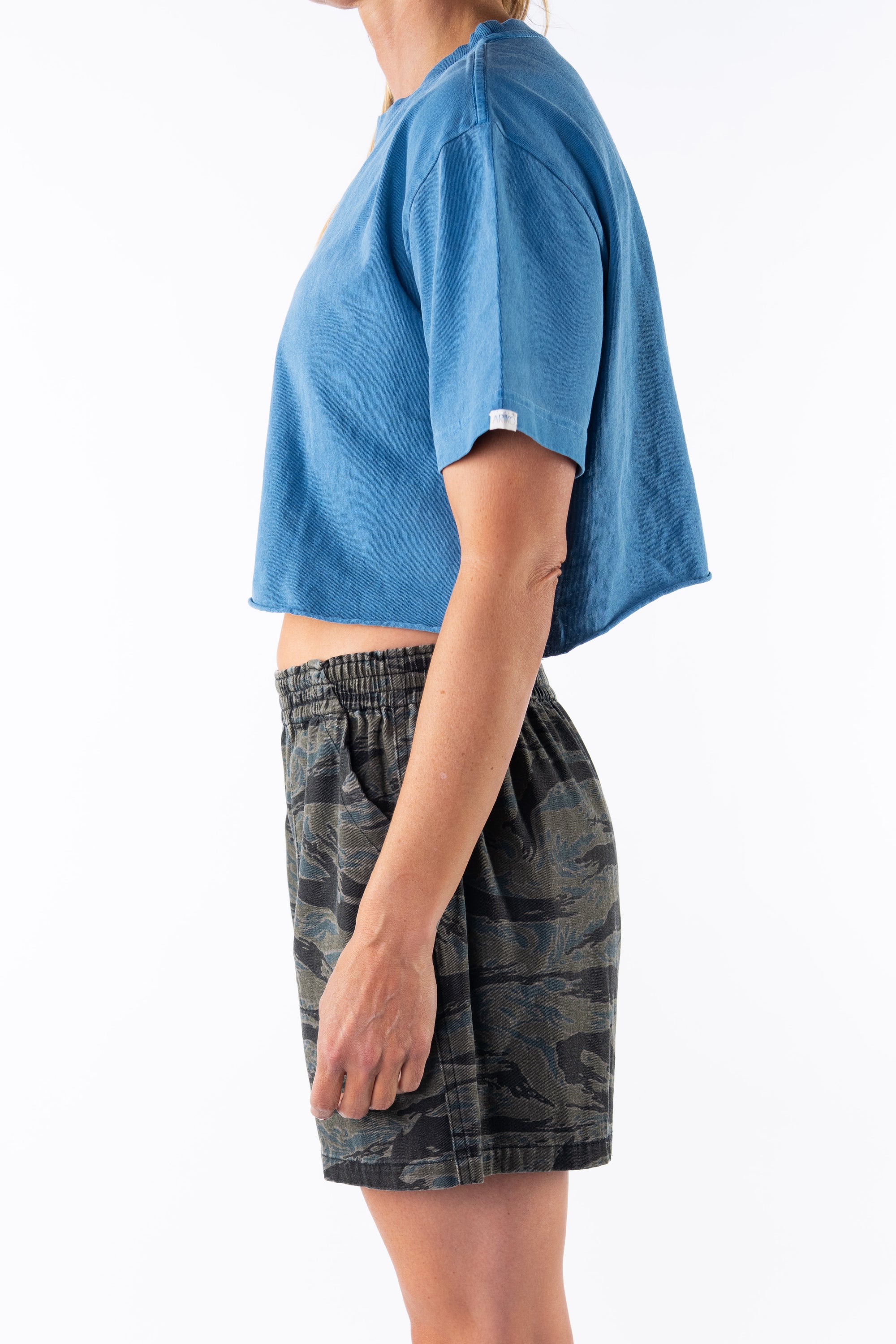 Side view of a woman wearing a teal crop t-shirt and ocean tiger camo day shorts on a white background