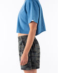 Side view of a woman wearing a teal crop t-shirt and ocean tiger camo day shorts on a white background