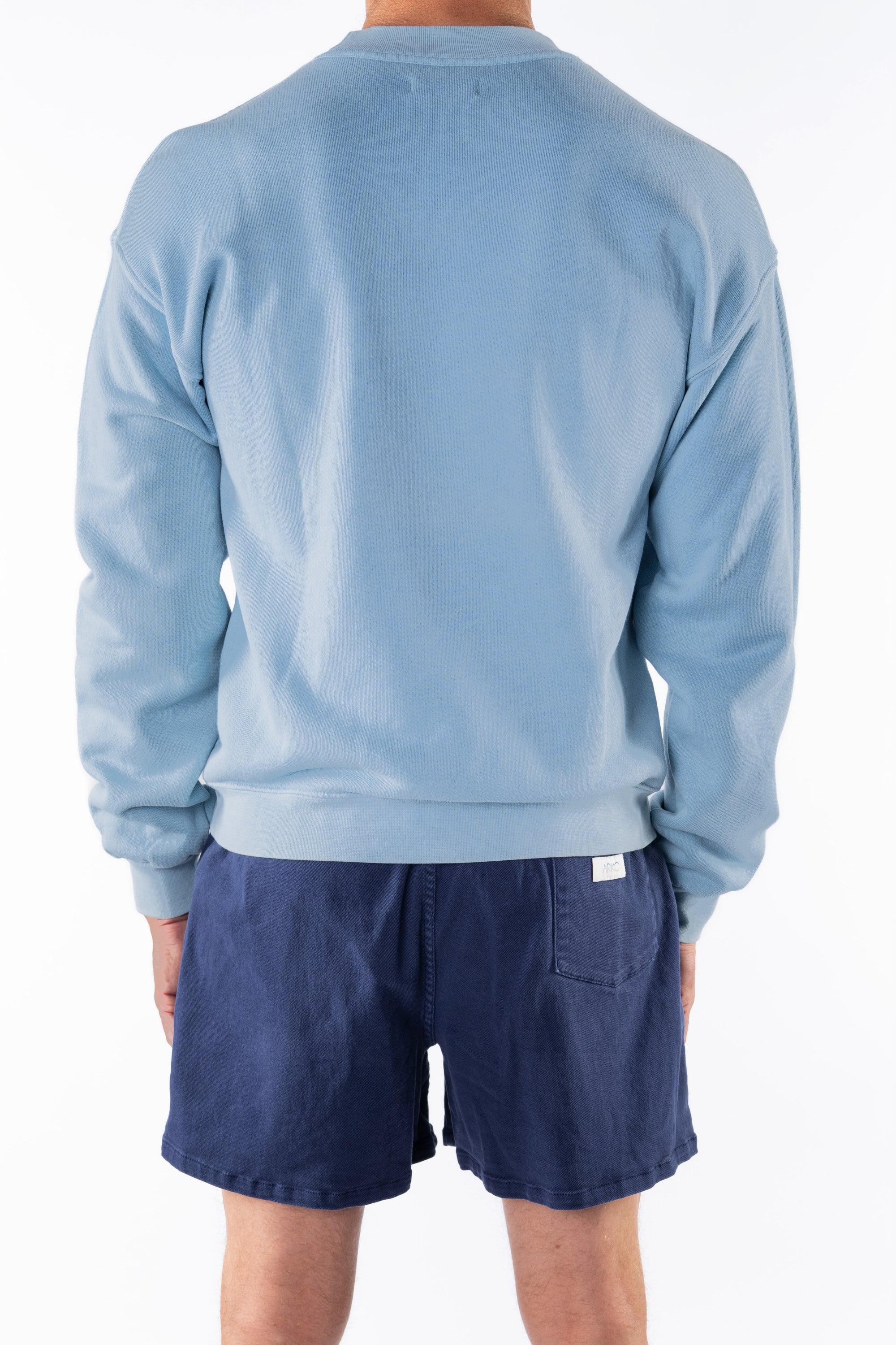 Back view of a person wearing a light blue ocean sweatshirt and navy day shorts on a white background