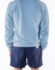 Back view of a person wearing a light blue ocean sweatshirt and navy day shorts on a white background