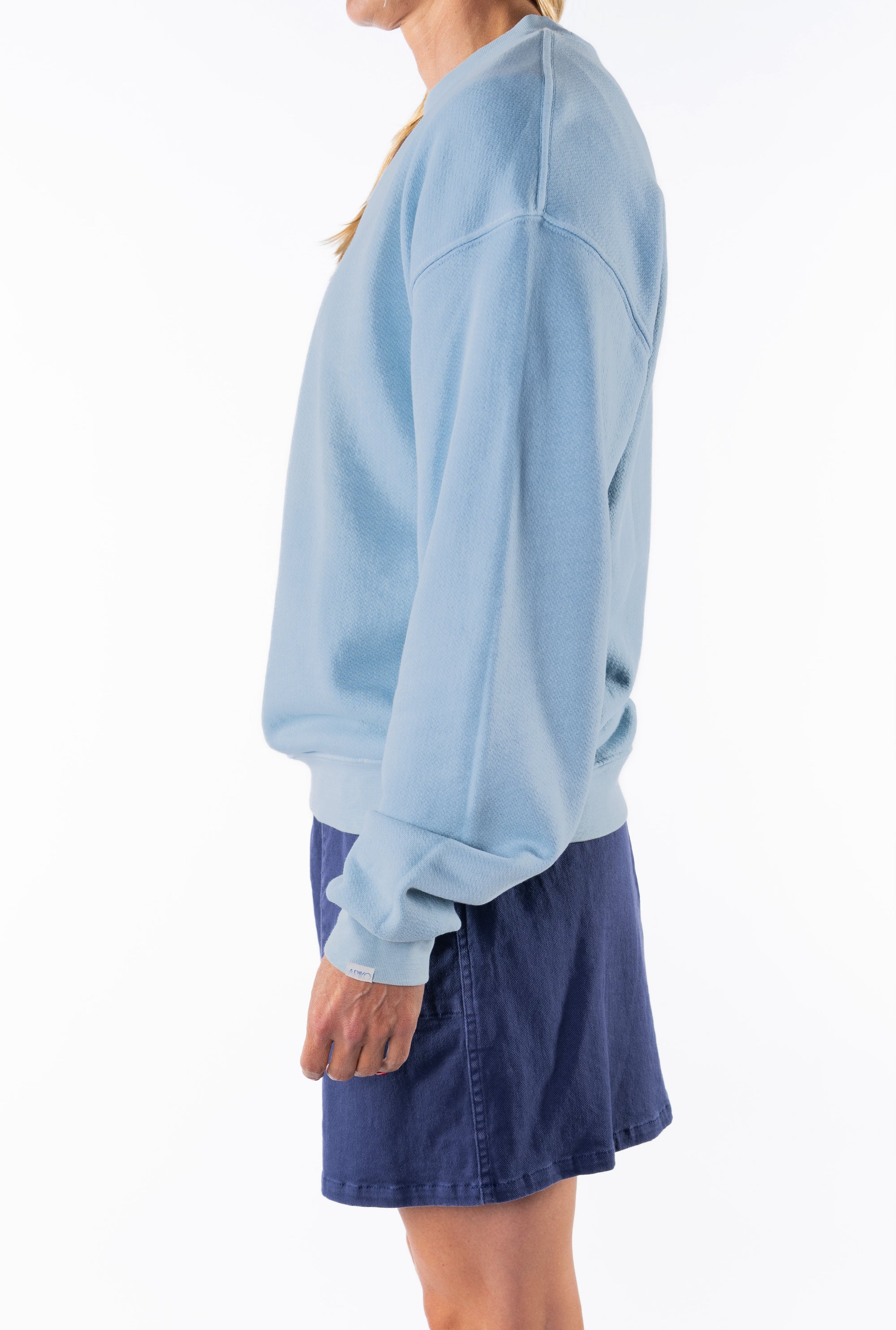 Side view of a woman wearing a light blue ocean sweatshirt and navy day skirt on a white background