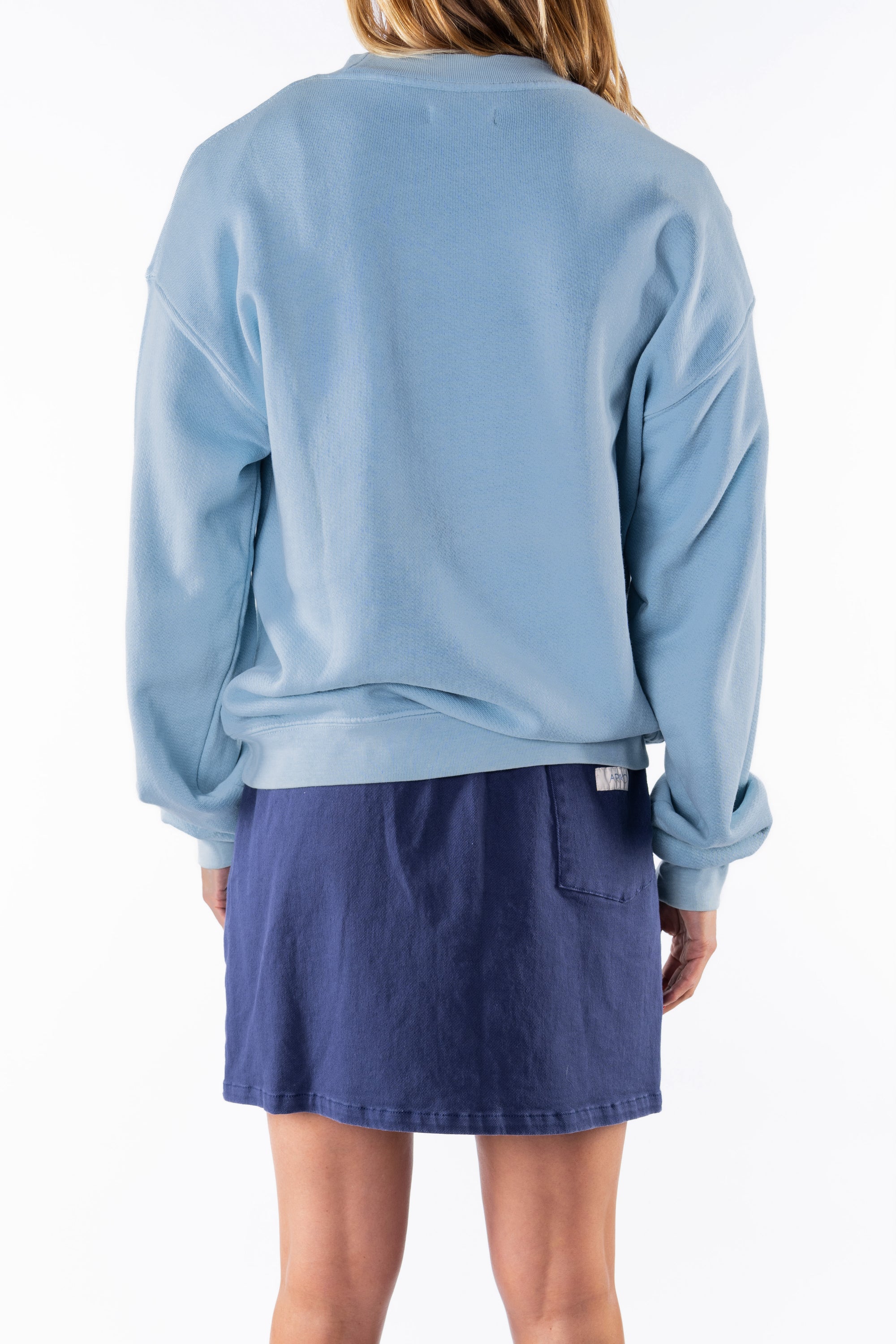 Back view of a woman wearing a light blue ocean sweatshirt and navy day skirt on a white background