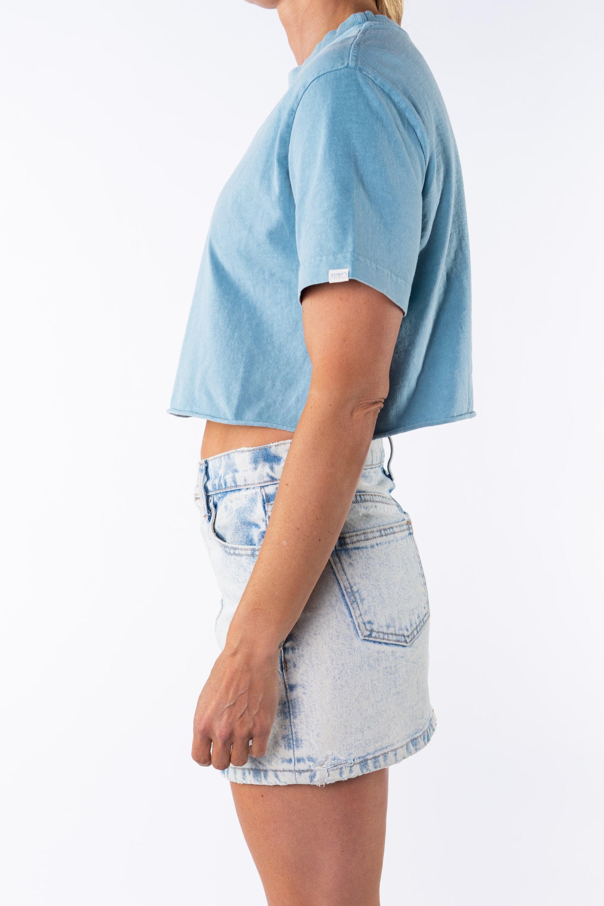 Side view of a woman wearing a turquoise beach crop t-shirt with a denim mini skirt on a white background. 