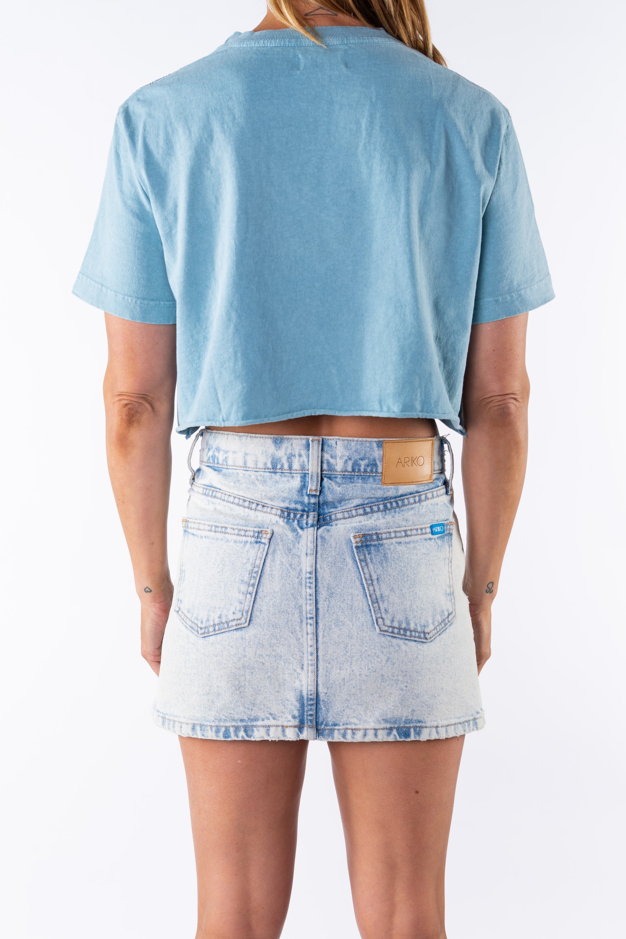 Back view of a woman wearing a light blue beach crop t-shirt and denim mini skirt on a white background
