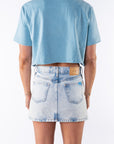 Back view of a woman wearing a light blue beach crop t-shirt and denim mini skirt on a white background